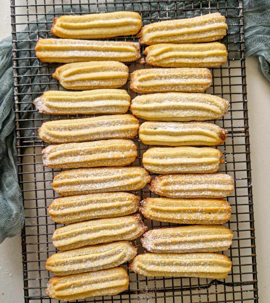 Prima imagine: Biscuiți lungi, dungați, aurii, dispuși pe un grătar de răcire, presărați ușor cu zahăr pudră. Aluatul lor bine definit păstrează forma în dungi datorită folosirii unui poș cu dui mare.