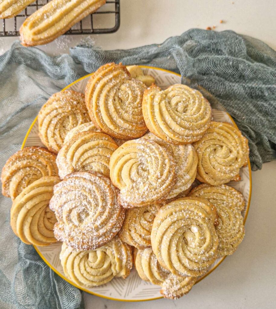 A doua imagine: Biscuiți spiralati, aurii și crocanți, stivuiți pe o farfurie rotundă, decorați cu zahăr pudră. Biscuiții au un aspect elegant datorită formei spiralate obținute cu un poș.