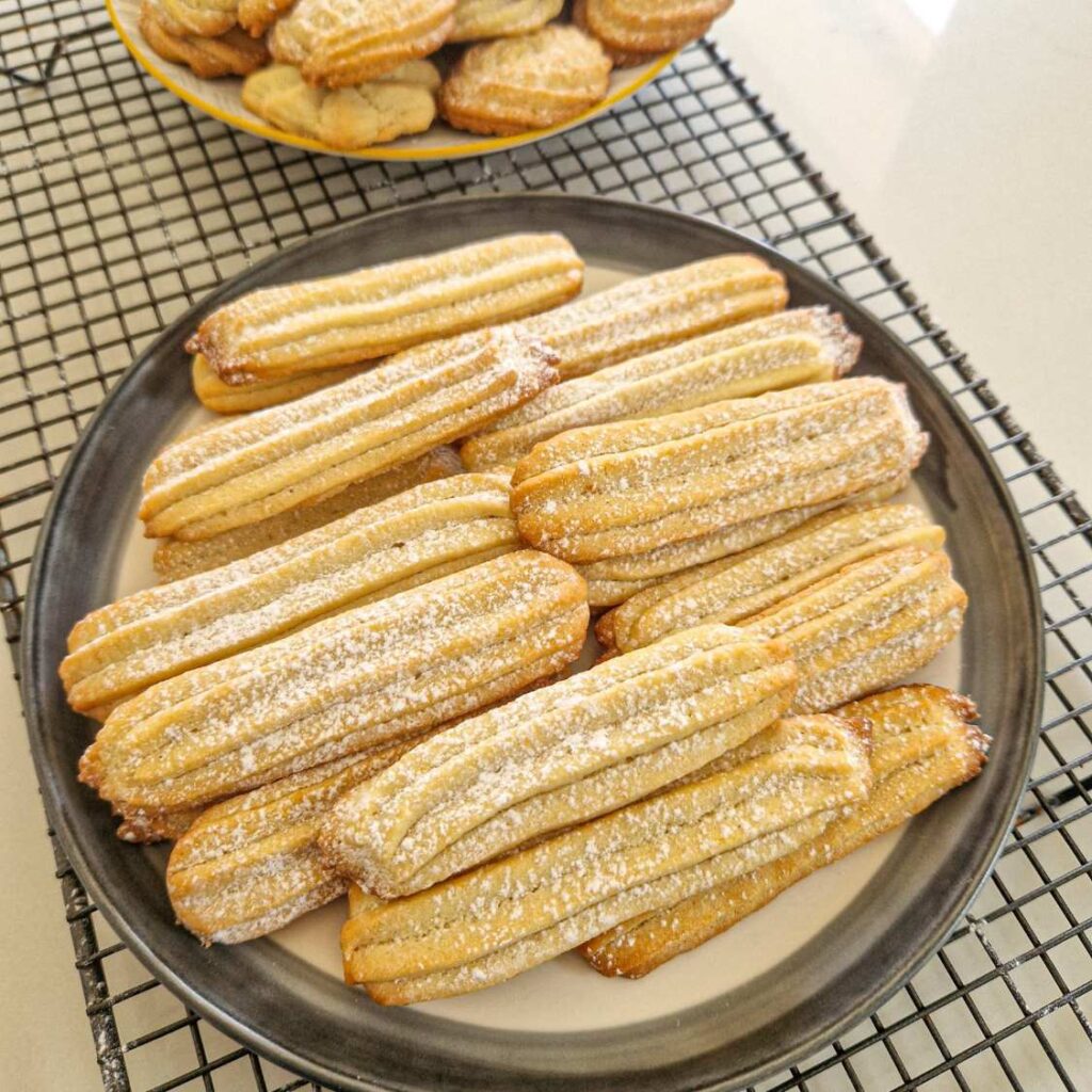 A treia imagine: Biscuiți lungi și aurii, puțin rumeniți pe margini, așezați pe o farfurie. Sunt presărați cu zahăr pudră, iar aluatul lor pare moale și bine definit.