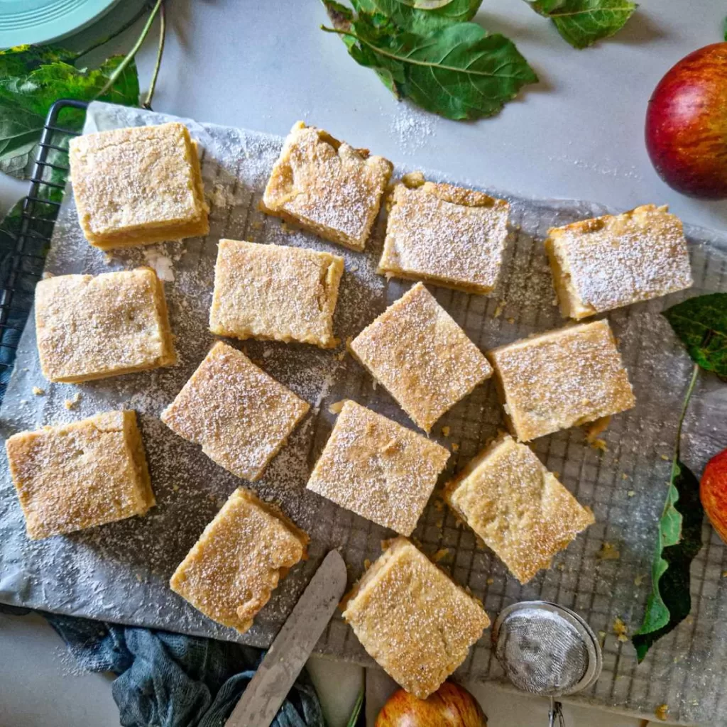 Prăjitură fragedă cu mere, tăiată în pătrate egale și presărată cu zahăr pudră, așezată pe hârtie de copt, într-o prezentare deasupra.