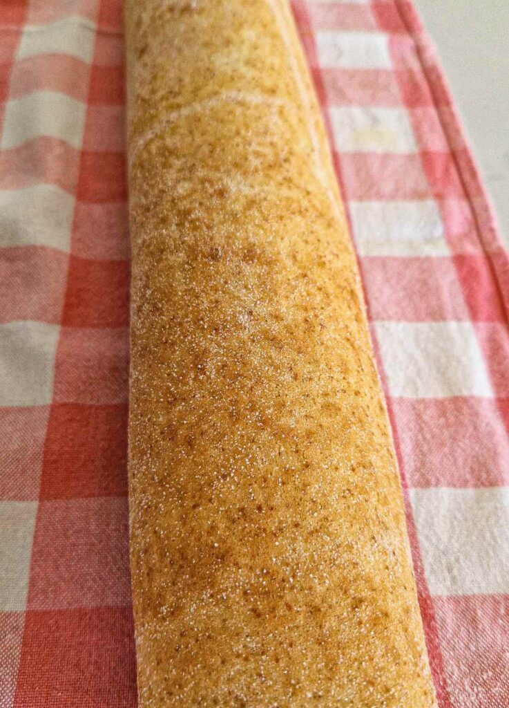 Rulada cu mere rulată și presărată cu zahăr, așezată pe un ștergar în carouri.