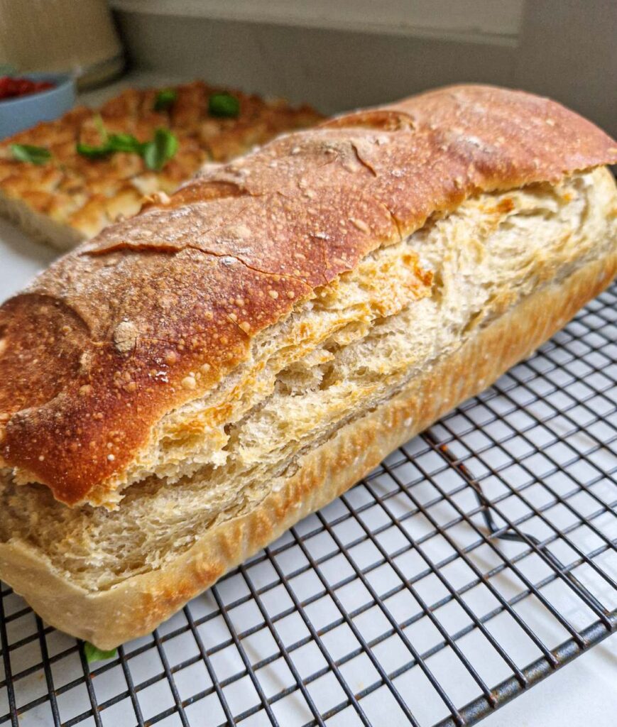 o paine coapta in tava, rumena aurie, unde se vede crusta crocanta si cat de frumos este crescuta, pe decor in spate este o focaccia decorata cu busuioc verde si rozmari, sare de mare