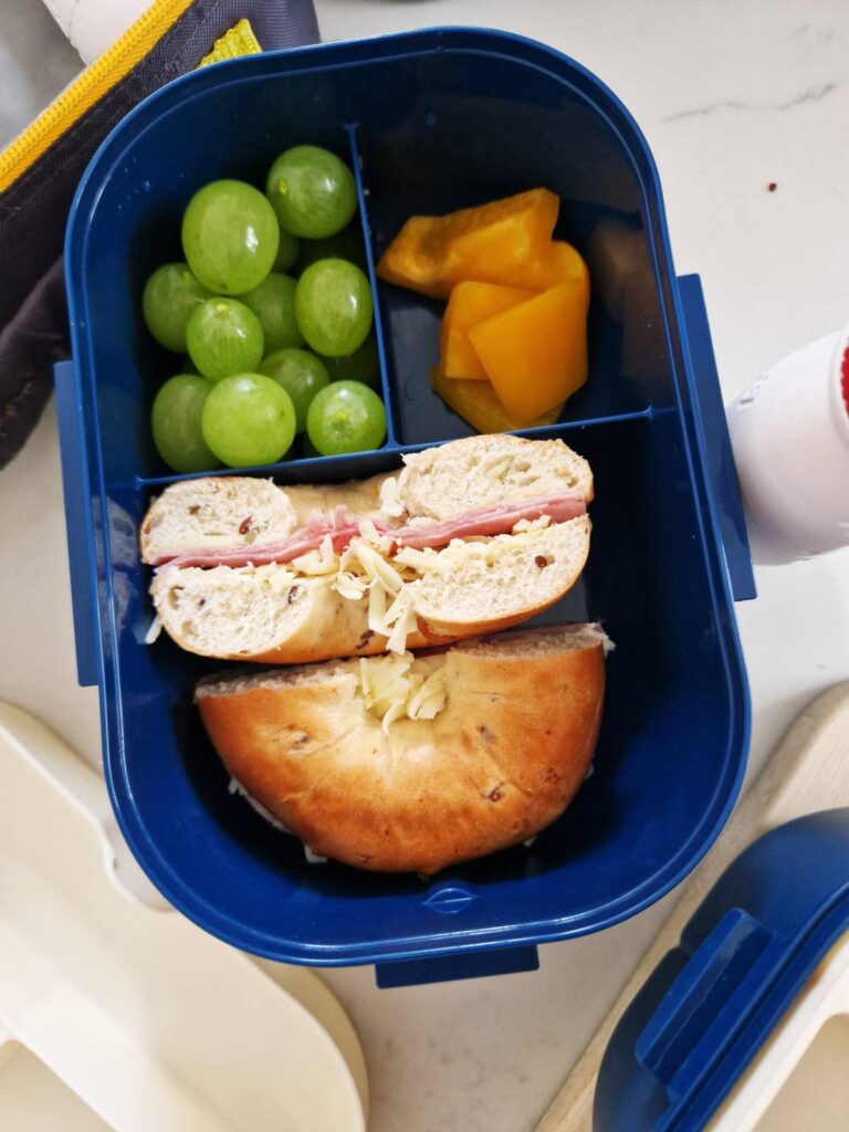 Lunchbox cu 3 compartimente: bagel pufos cu cașcaval și ham, tăiat pe jumătate, alături de boabe de struguri și bucățele de castravete proaspăt