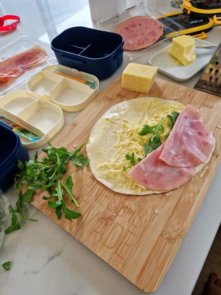 Masa de lucru incarcata cu toatecele necesare pentru pregatirea pachetelului cu gust , wrap , cascaval , rucola, cutia de impachetat 