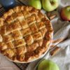 Apple Pie cu unt – rețeta clasică, pas cu pas - aurie abia scoasadin cuptor, cu un decor rustic cu prosop, baton de scortisoara, mere pe un gratar