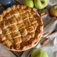Apple Pie cu unt – rețeta clasică, pas cu pas - aurie abia scoasadin cuptor, cu un decor rustic cu prosop, baton de scortisoara, mere pe un gratar