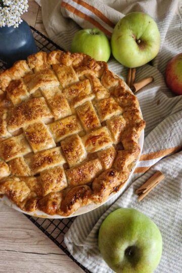 Apple Pie cu unt – rețeta clasică, pas cu pas - aurie abia scoasadin cuptor, cu un decor rustic cu prosop, baton de scortisoara, mere pe un gratar