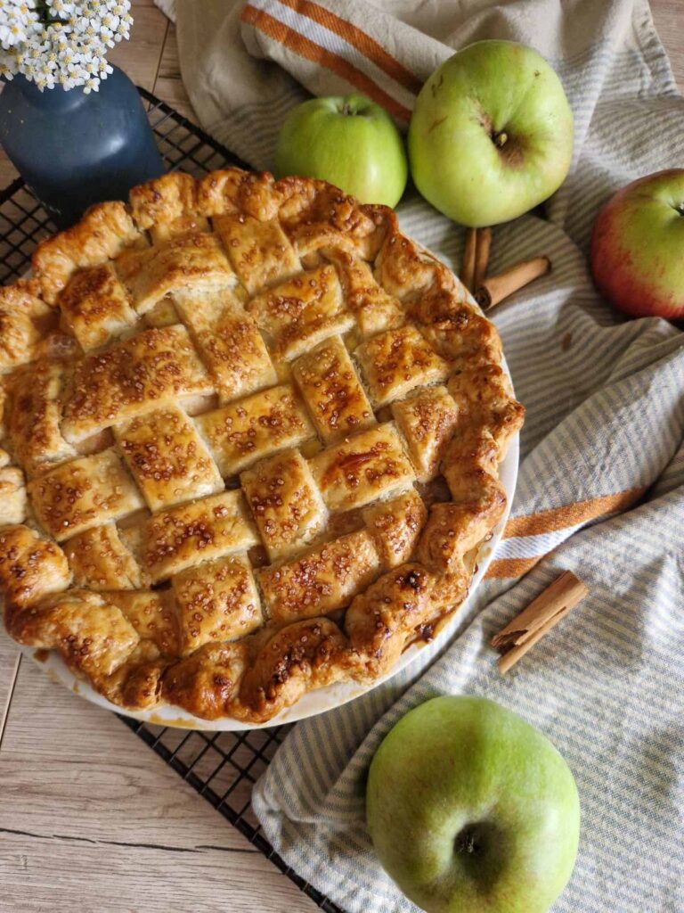 Apple Pie cu unt – rețeta clasică, pas cu pas - aurie abia scoasadin cuptor, cu un decor rustic cu prosop, baton de scortisoara, mere pe un gratar 