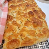 Focacia întreagă, așezată pe masă, cu crustă aurie și frunze de rozmarin, văzută de la distanță.