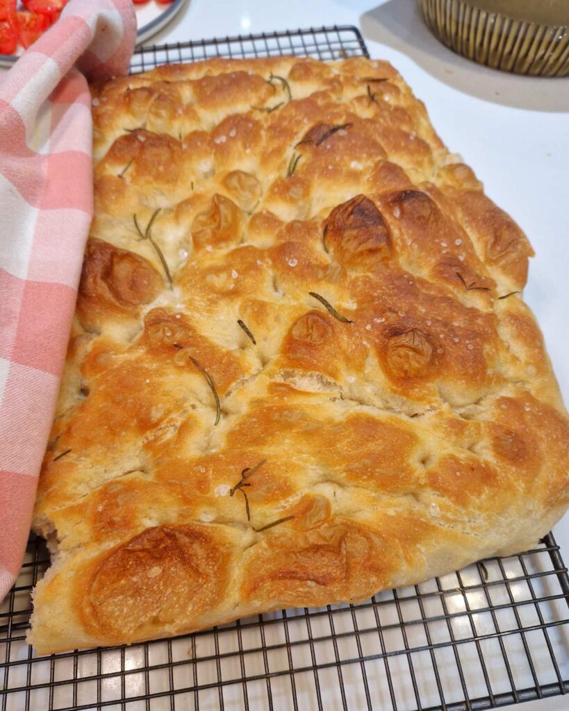 Focacia întreagă, așezată pe masă, cu crustă aurie și frunze de rozmarin, văzută de la distanță.