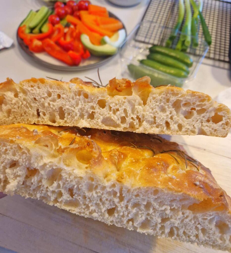 Focacia tăiată pe jumătate, văzută din unghi care arată ambele părți, cu textura aerată și bulgări crocanți pe suprafață, decorată cu rozmarin.