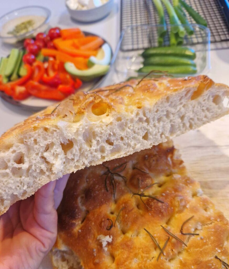 Focacia mare tăiată pe jumătate, cu o crustă aurie și interior pufos, presărată cu rozmarin proaspăt.