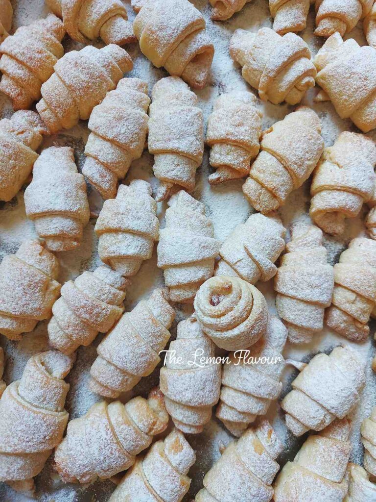 Cornulețe fragede cu untură și smântână, toate împudrate cu zahăr vanilat, așezate într-o tavă mare, gata de servire.