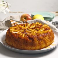 Prajitura Rasturnata cu mere pe platou, alaturi de un decor cu flori, mere si farfurii si furculite gata de servit