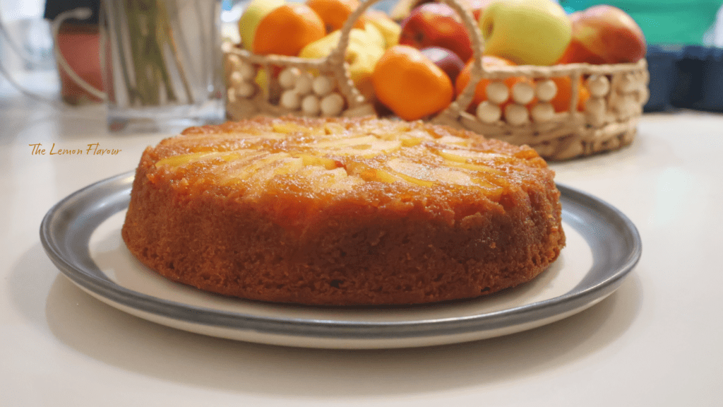 Prajitura Rasturnata cu mere pe platou, alaturi de un decor cu flori, si uncoscu fructe, unde se var merele caramelizate aurii